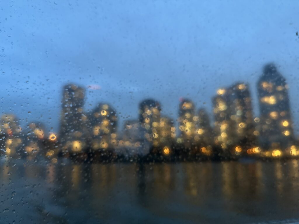 Blurry buildings of Vancouver's downtown at night -- the focus is on the rain drops across the boat's windows