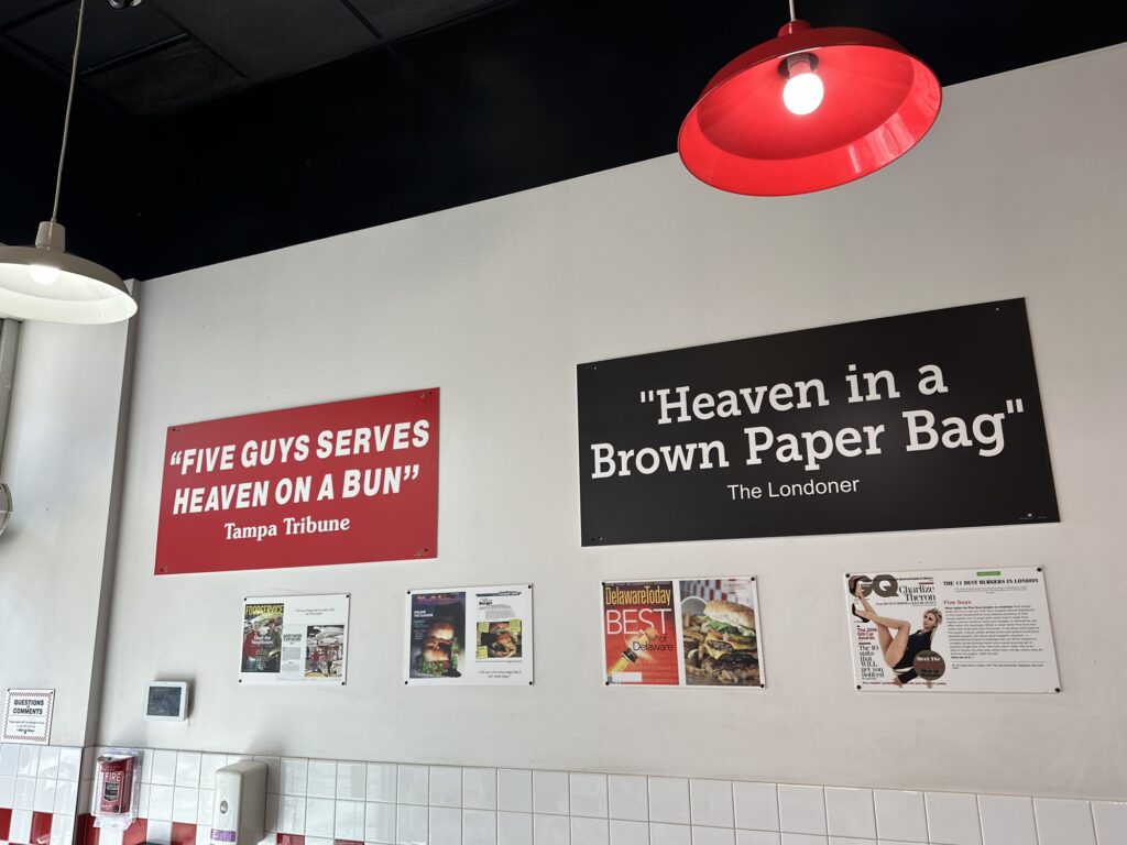 A photo from inside a "Five Guys" burger joint, with some of the press quotes praising the restaurant. One quote says "Five Guys serves heaven on a bun" while the other quote says "Heaven in a brown paper bag".