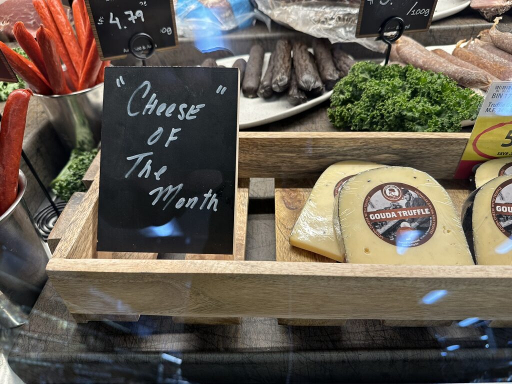 A Deli counter at a supermarket with some cheese, and a sign that says “”Cheese” of the month”, and yes the word Cheese is in quotes on the sign…