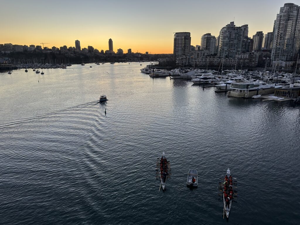A photo of False Creek, with downtown Vancouver on the right, and Kitsilano on the left. Some dragonboats are preparing to race. A little ferry boat is cruising across the water. The photo is taken from the Cambie bridge.
