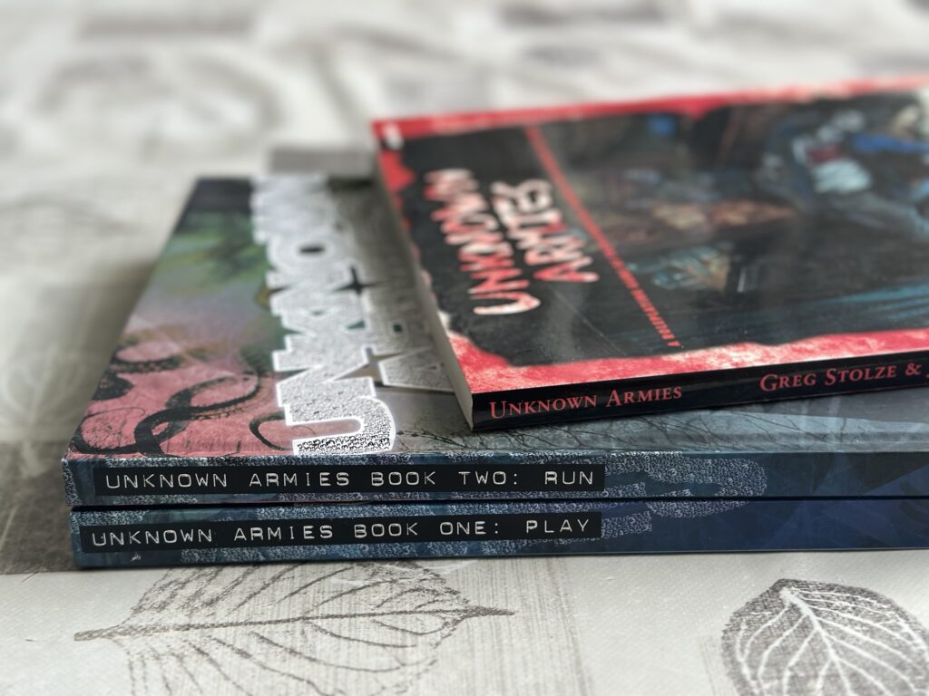 Unknown Armies first edition on top of the 3rd edition's Play and Run books.